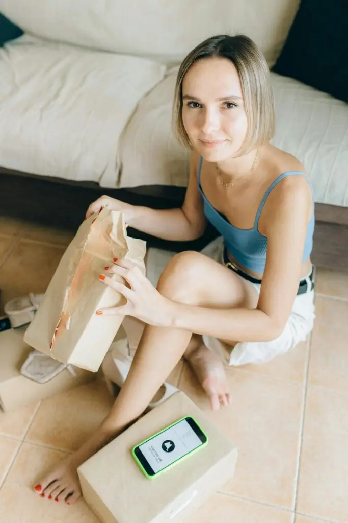A young woman sits in her living room, opening a package with a smartphone nearby.